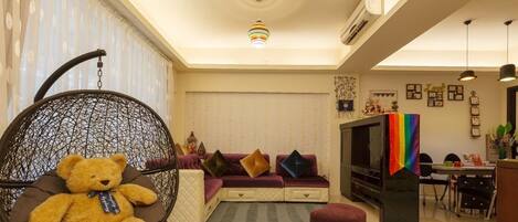 A living room with a sofa, a hanging chair with a teddy bear, a TV, and a dining area.