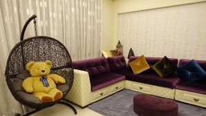 Una habitación con un sofá morado grande, una silla colgante con un osito de peluche y un pequeño reposapiés.