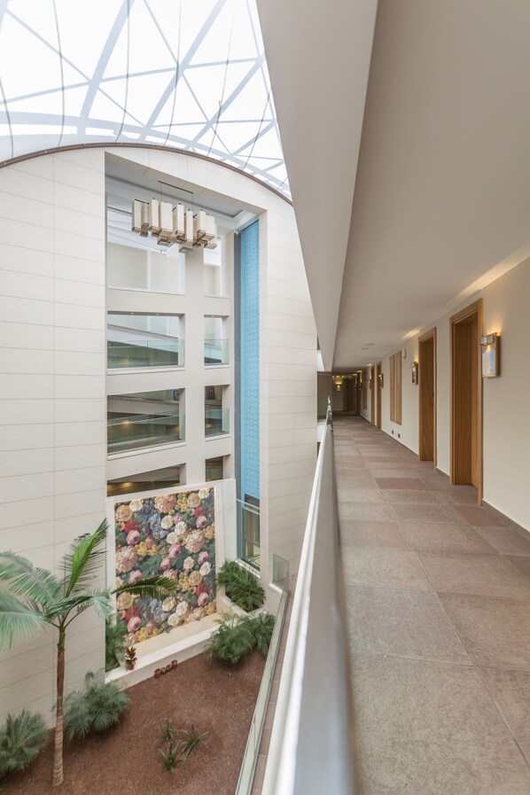 A modern hotel corridor with a glass ceiling, a floral wall mural, and a view of the sky.
