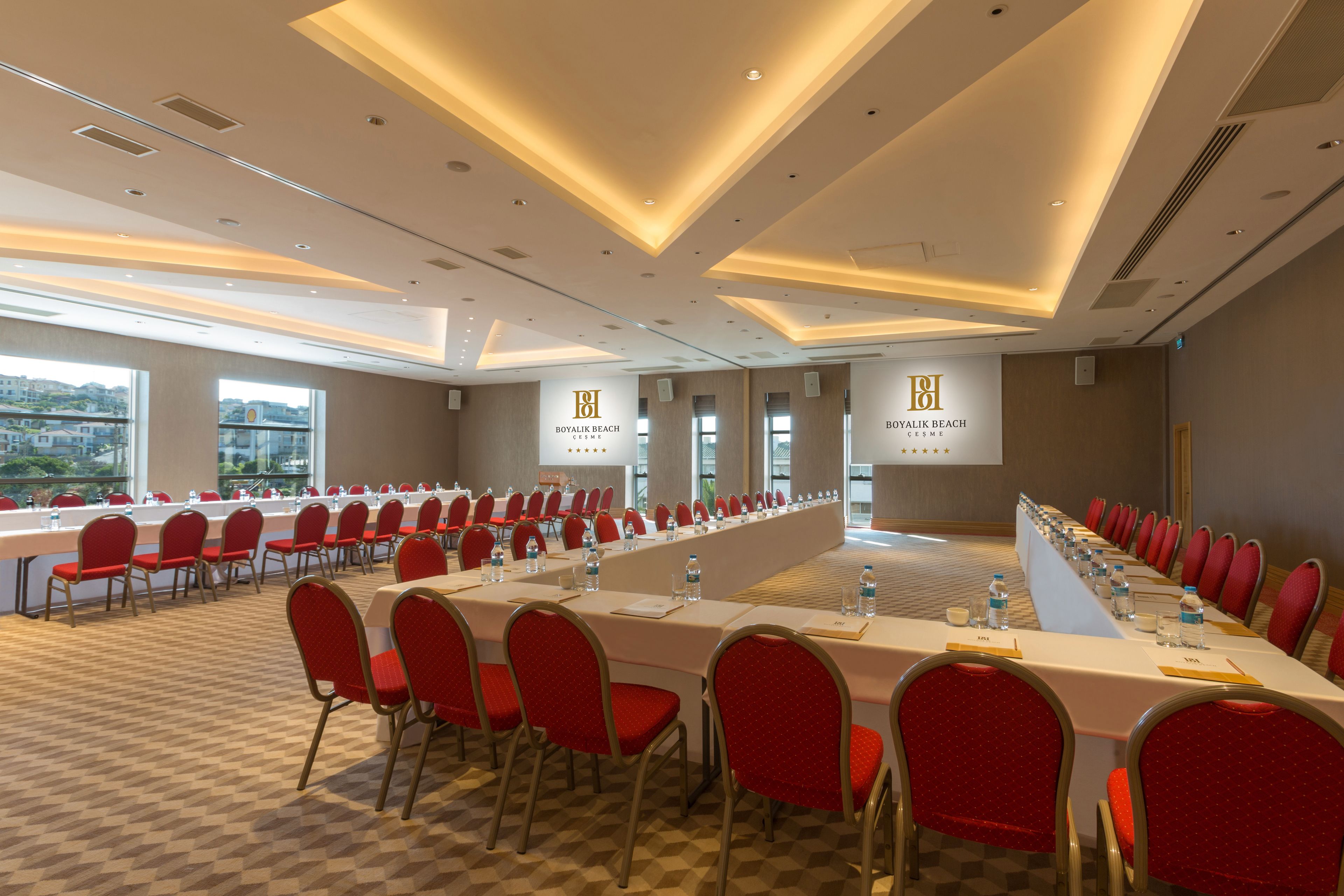 A conference room with a long table, red chairs, and a view of a cityscape through large windows.