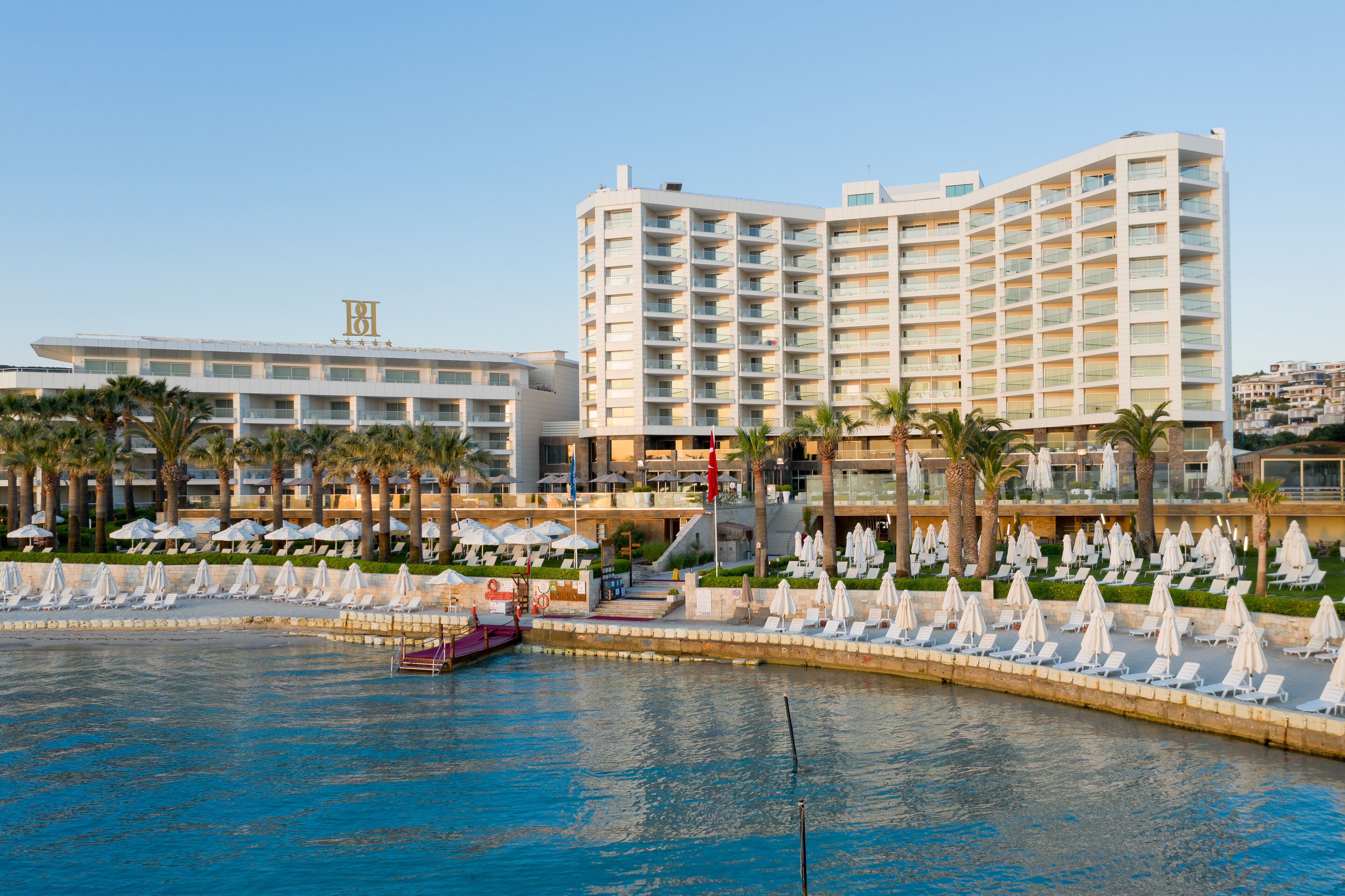 Ein Hotel mit mehreren Etagen, einem Poolbereich inklusive Liegestühle und Sonnenschirme sowie einer Wasserfläche im Vordergrund.
