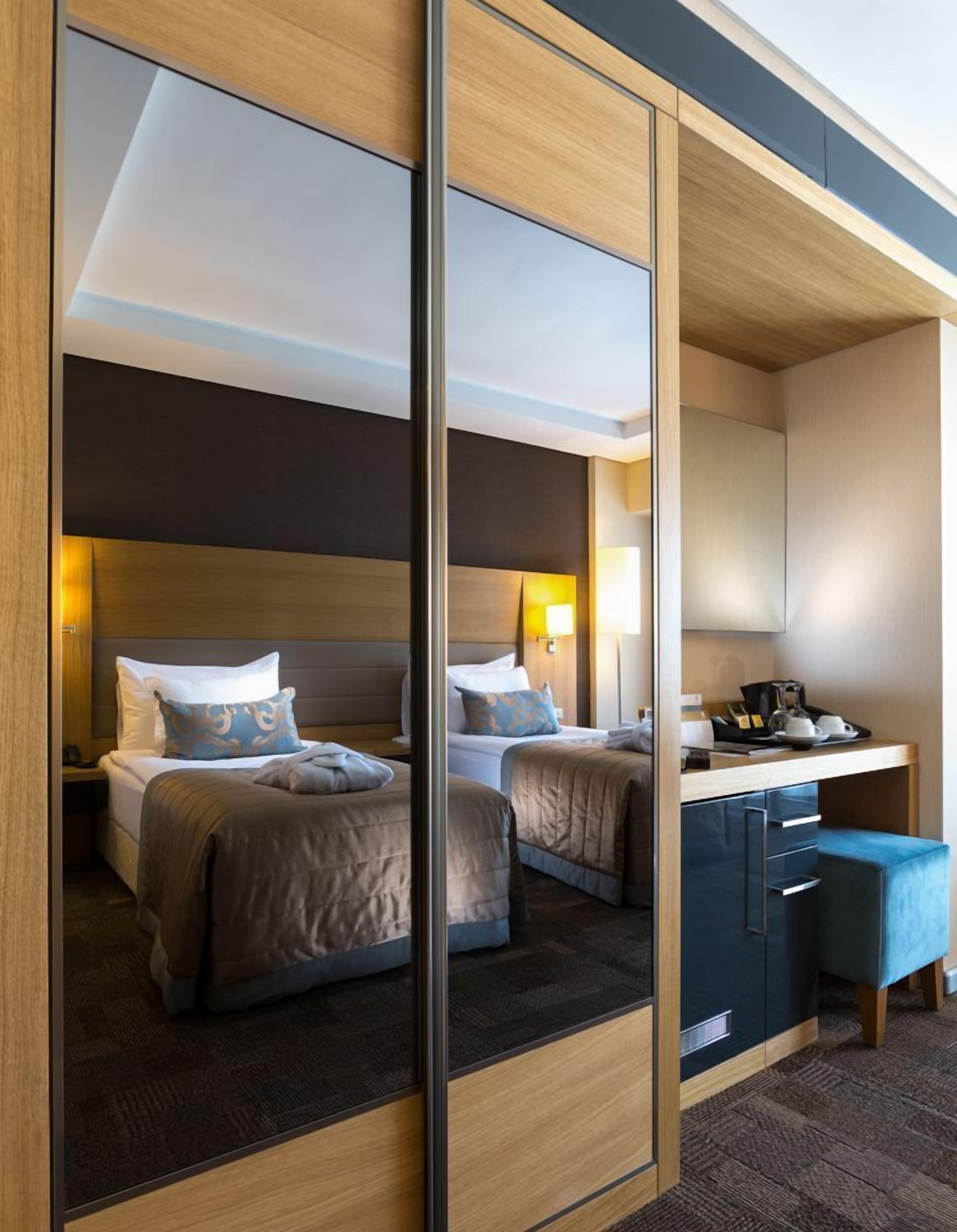A modern hotel room with a large mirror, two beds, a desk with a chair, and a small seating area.