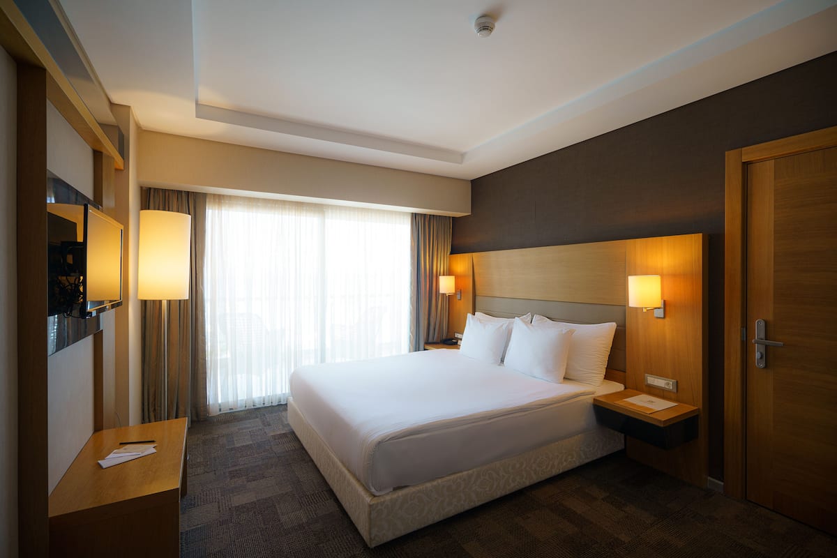 Quarto de hotel com uma cama grande, duas mesas de cabeceira, uma escrivaninha, uma televisão e uma janela com cortinas.