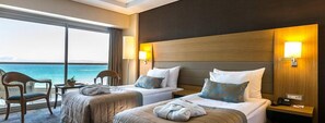 A hotel room with two beds, a large window with a view of the sea, and a small table with a chair.