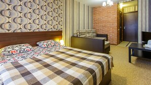 A hotel room with a checkered bedspread, a patterned headboard, a sofa with a plaid cushion, a small table, and a brick accent wall.