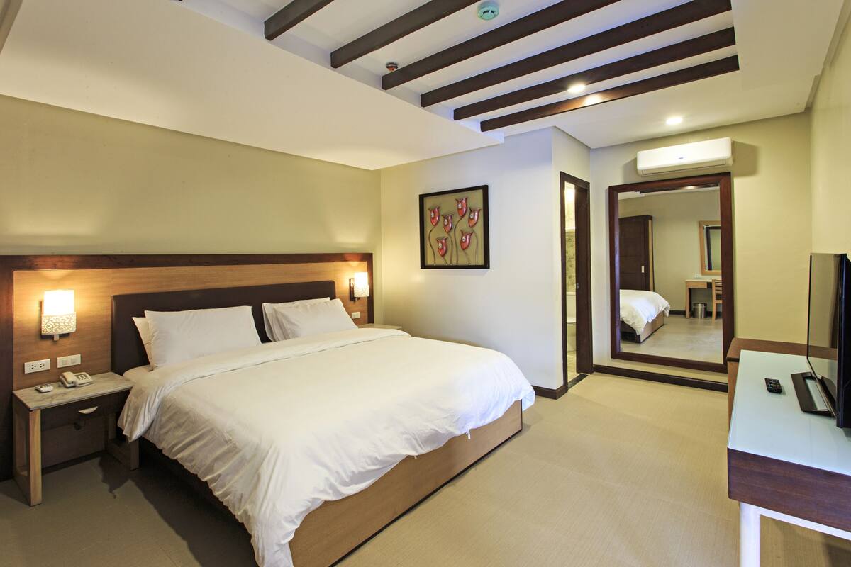 Ein Hotelzimmer mit einem großen Bett, Nachttischen, einem Schreibtisch, einem Spiegel und einer Decke mit Holzbalken.