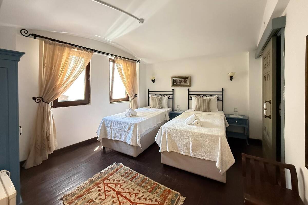 A bedroom with two beds, a window with curtains, a bedside table with a lamp, and a patterned rug.