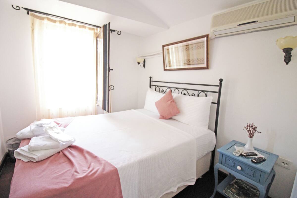 A neatly made bed with a white and pink bedspread, a small bedside table with a vase and remote controls, and a window with curtains.