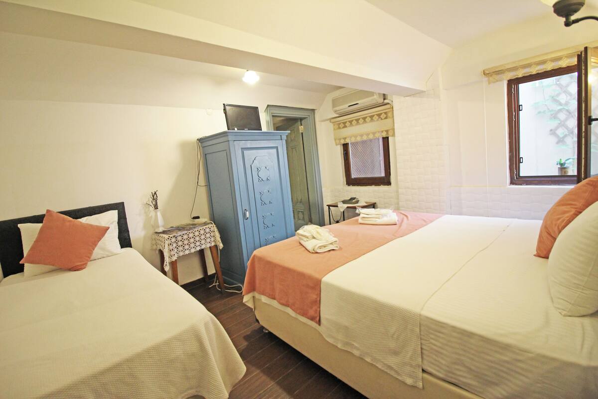 A bedroom with two beds, a small table, a cabinet, and a window with a view.