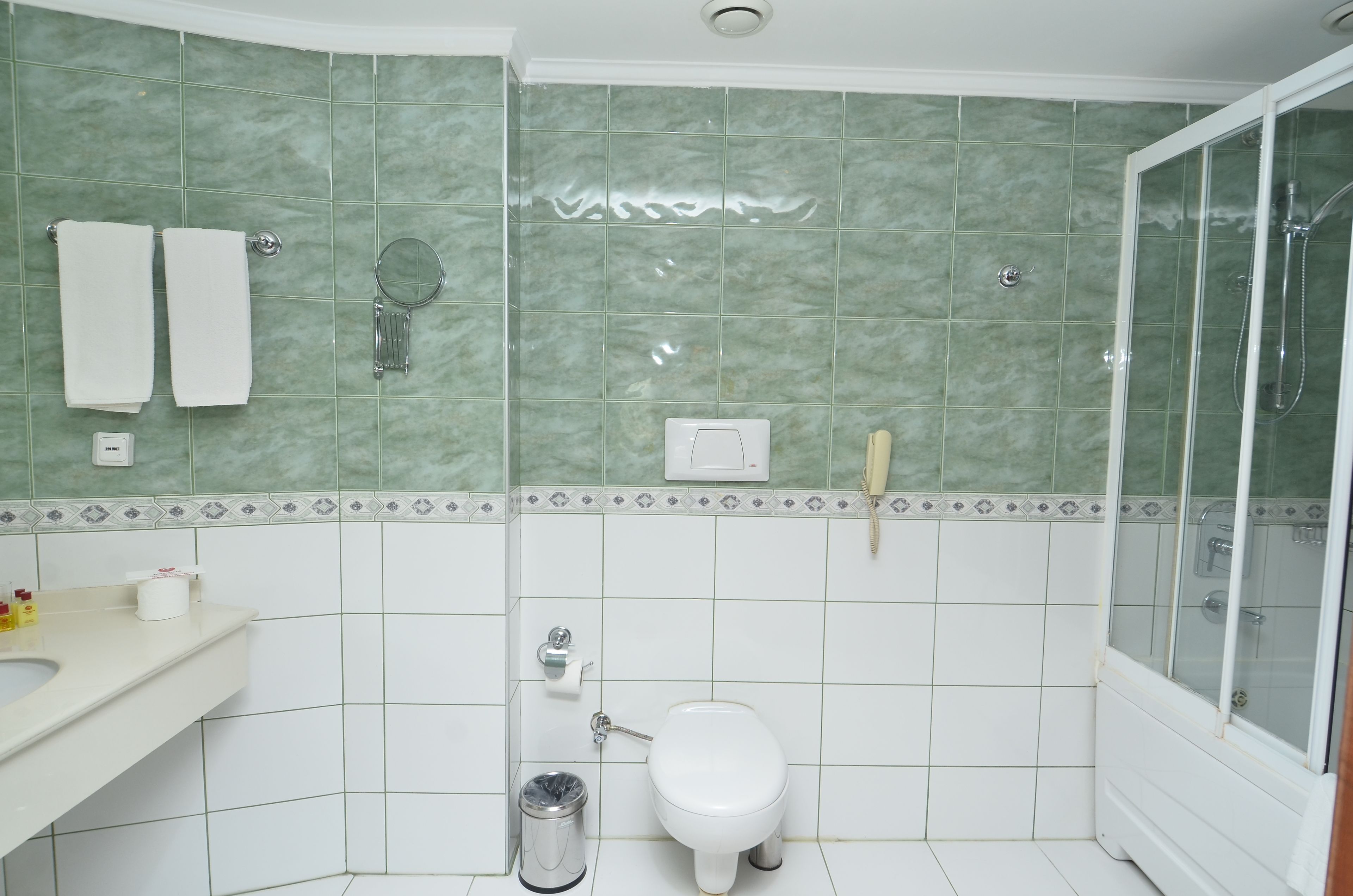 A bathroom with green tiles, a toilet, a sink, a trash bin, and a shower area.