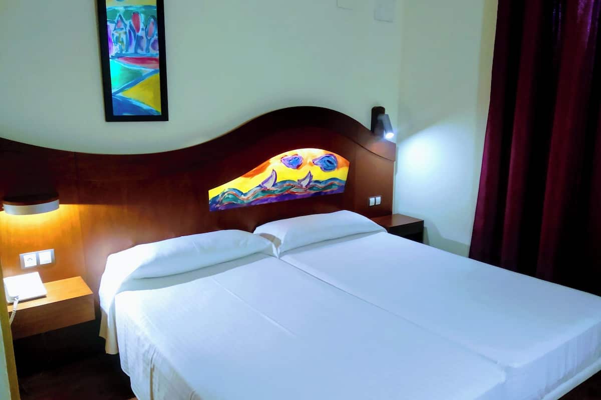 A hotel room with a double bed, a nightstand with a lamp, a painting on the headboard, and a window with curtains.