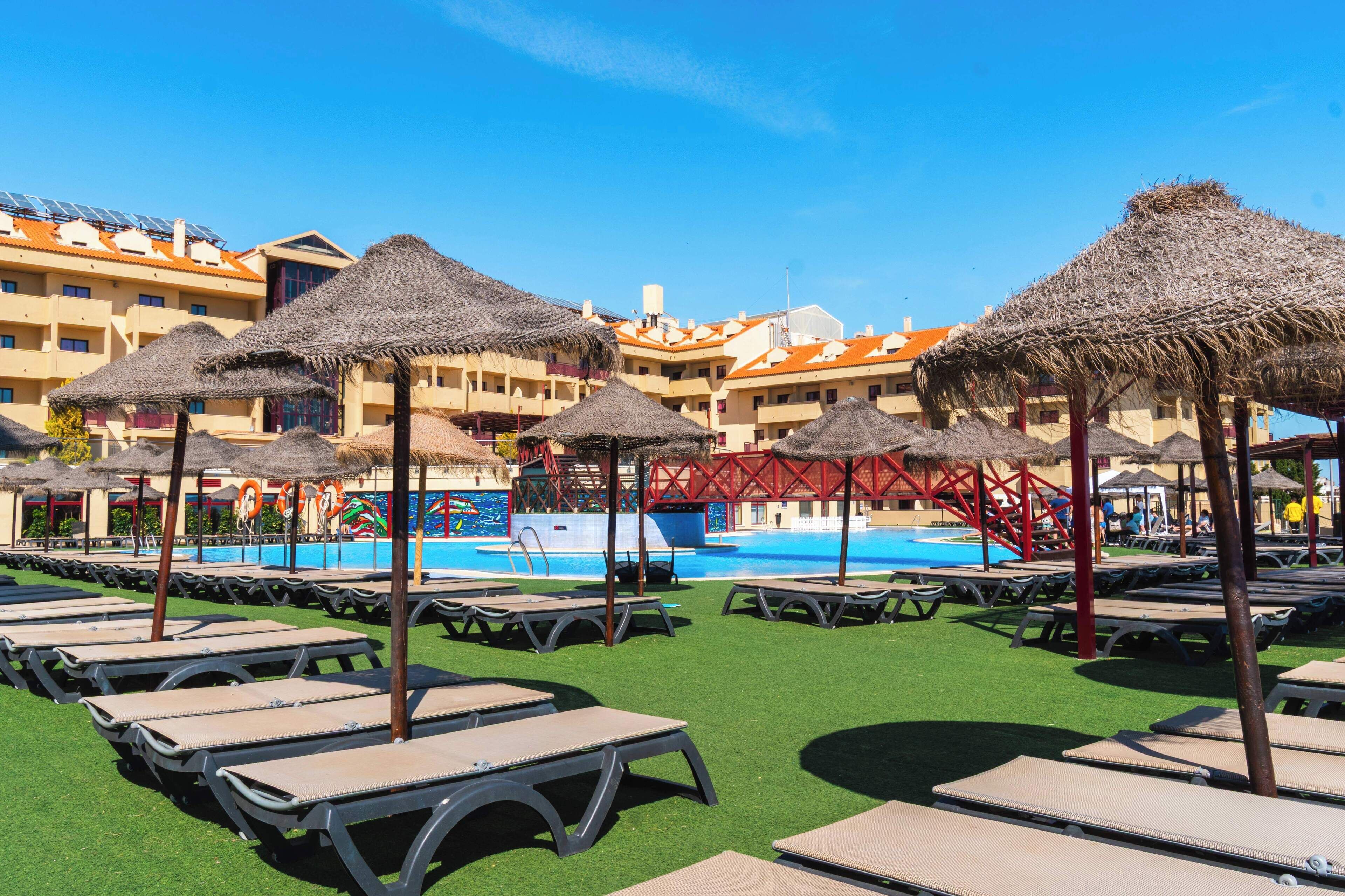 A poolside area with sun loungers and thatched umbrellas, a swimming pool, and a building with orange roofs in the background.