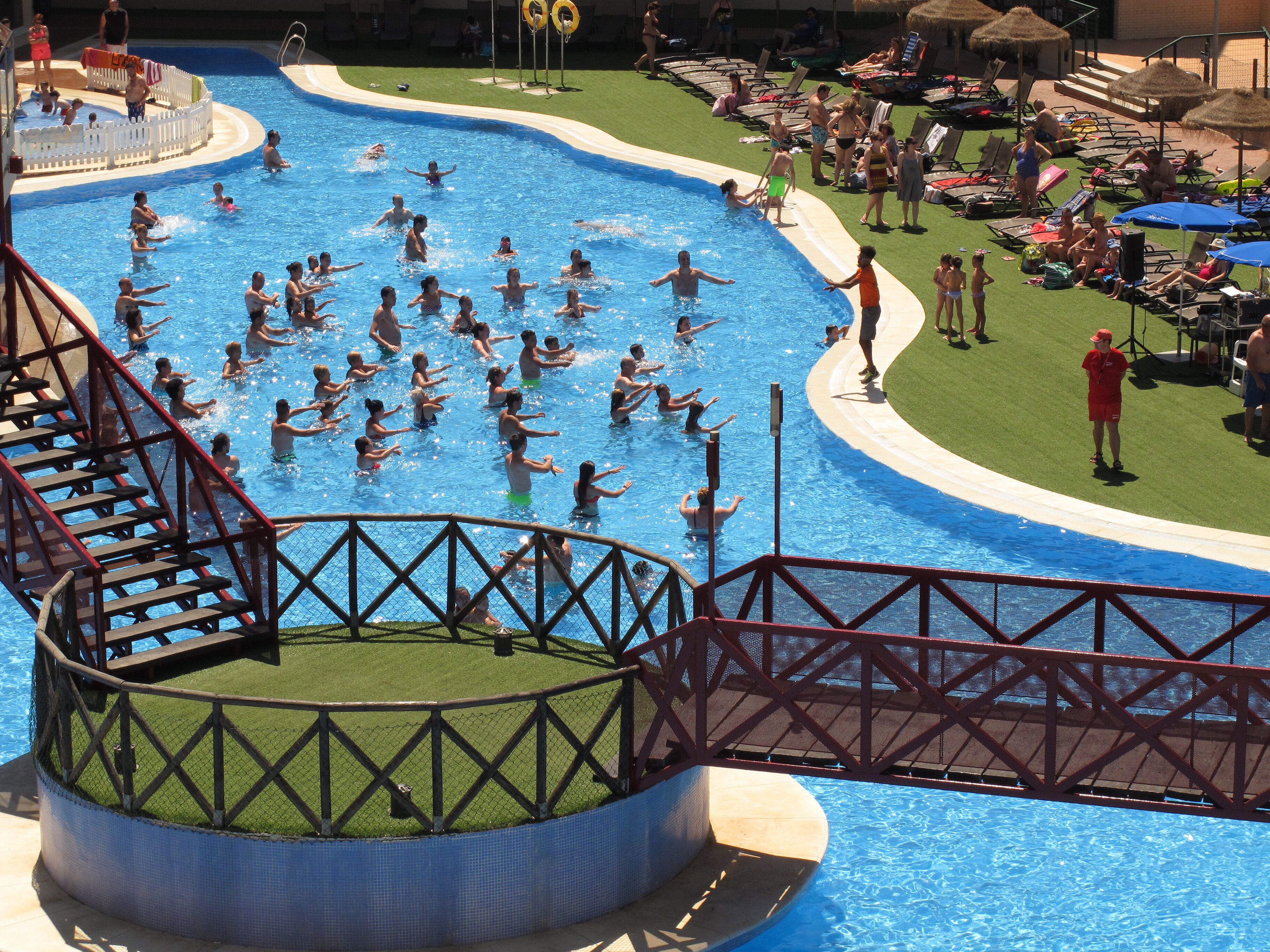 Una gran piscina con un área de terraza alrededor, una pequeña fuente circular y una zona vallada con un tobogán.