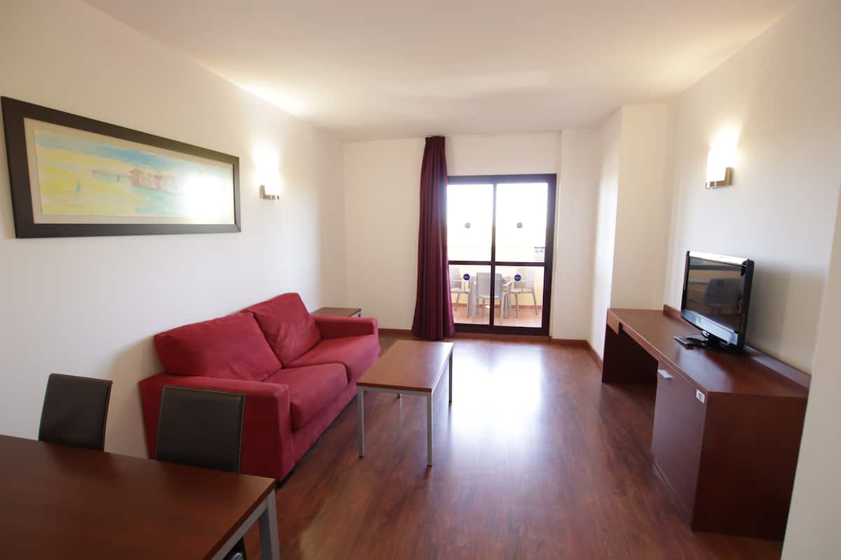 A living room with a red sofa, a wooden coffee table, a TV on a wooden stand, and a sliding glass door leading to a balcony.