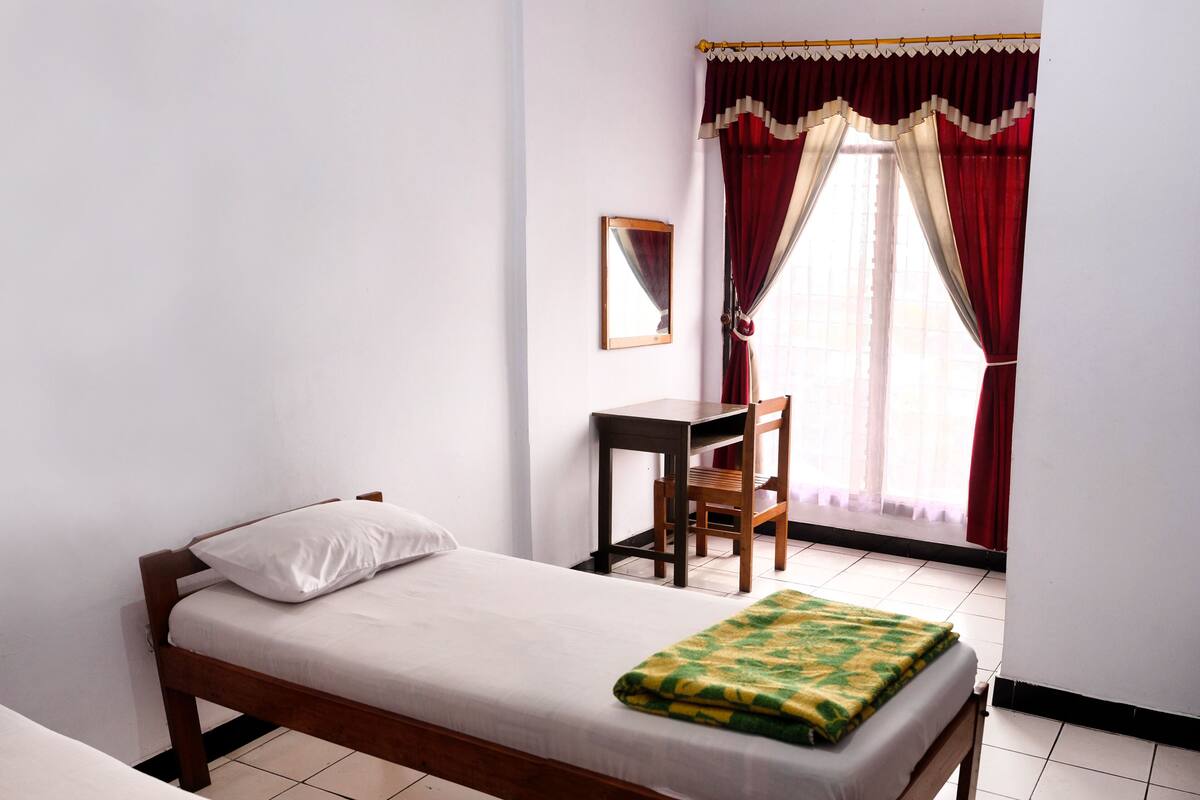 A single bed with a white mattress and a green and yellow blanket, a wooden chair, a small wooden table, and a window with red curtains.
