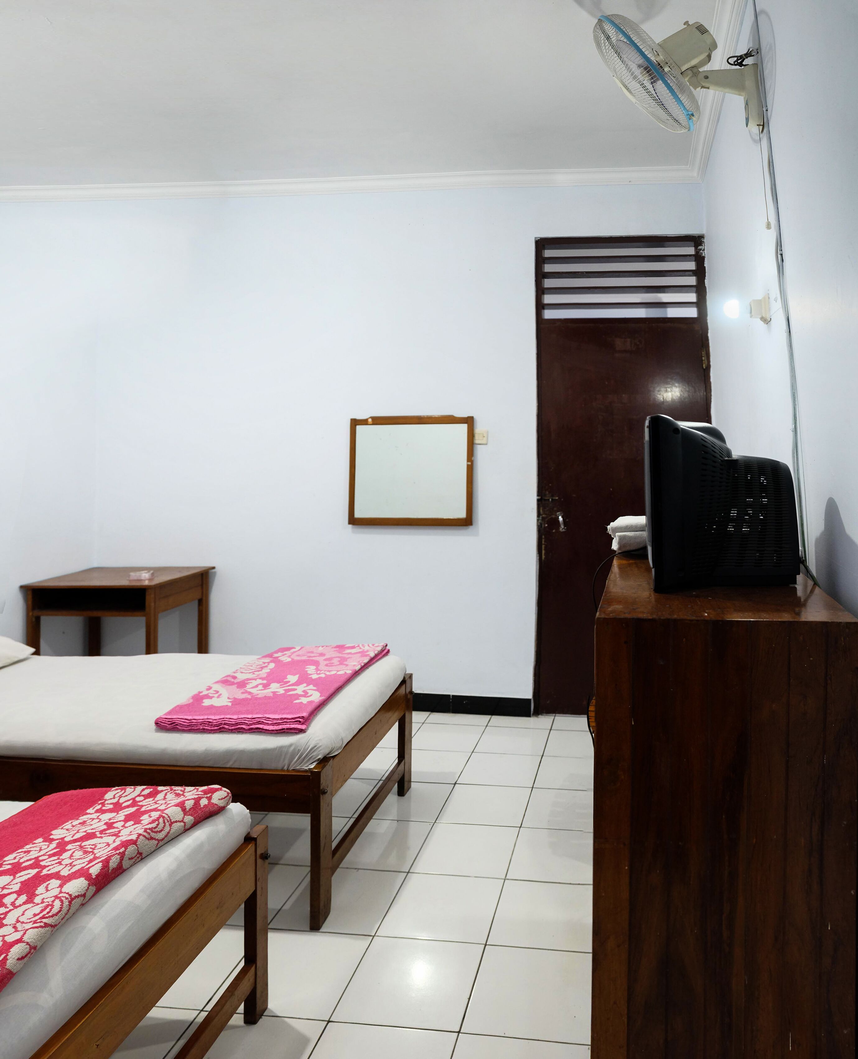 Quarto com duas camas de madeira, uma pequena mesa de madeira, uma escrivaninha com cadeira preta, um ventilador fixado na parede e uma porta com janela.
