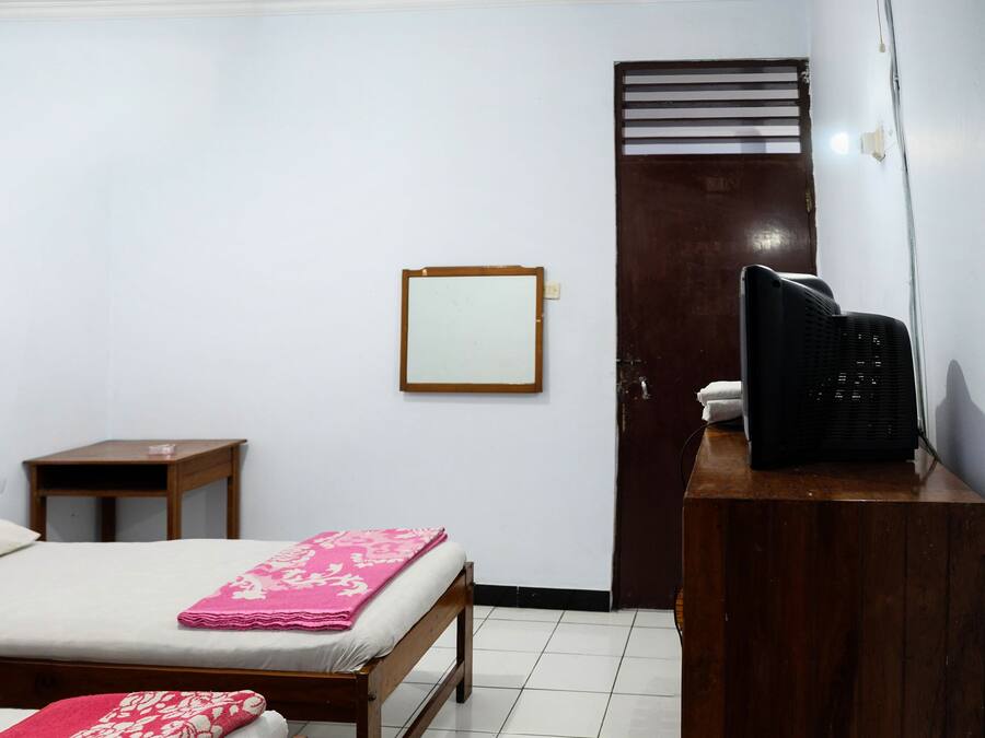 Quarto com duas camas de madeira, uma pequena mesa de madeira, uma escrivaninha com cadeira preta, um ventilador fixado na parede e uma porta com janela.
