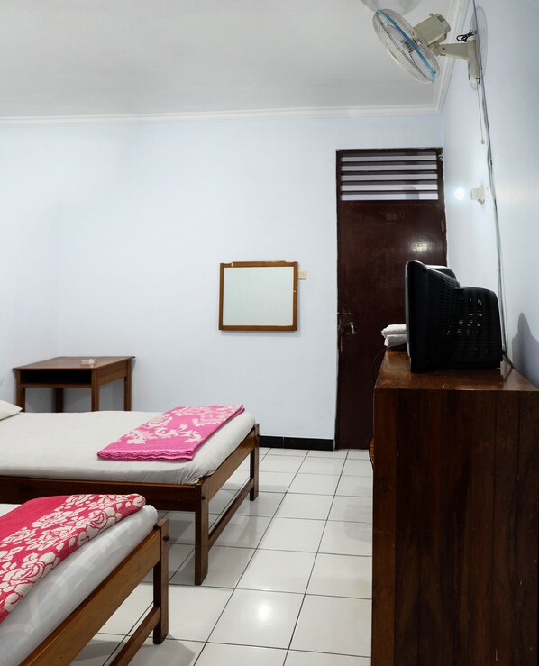A room with two wooden beds, a small wooden table, a desk with a black chair, a wall-mounted fan, and a door with a window. - Hotel Wisata (Magelang)