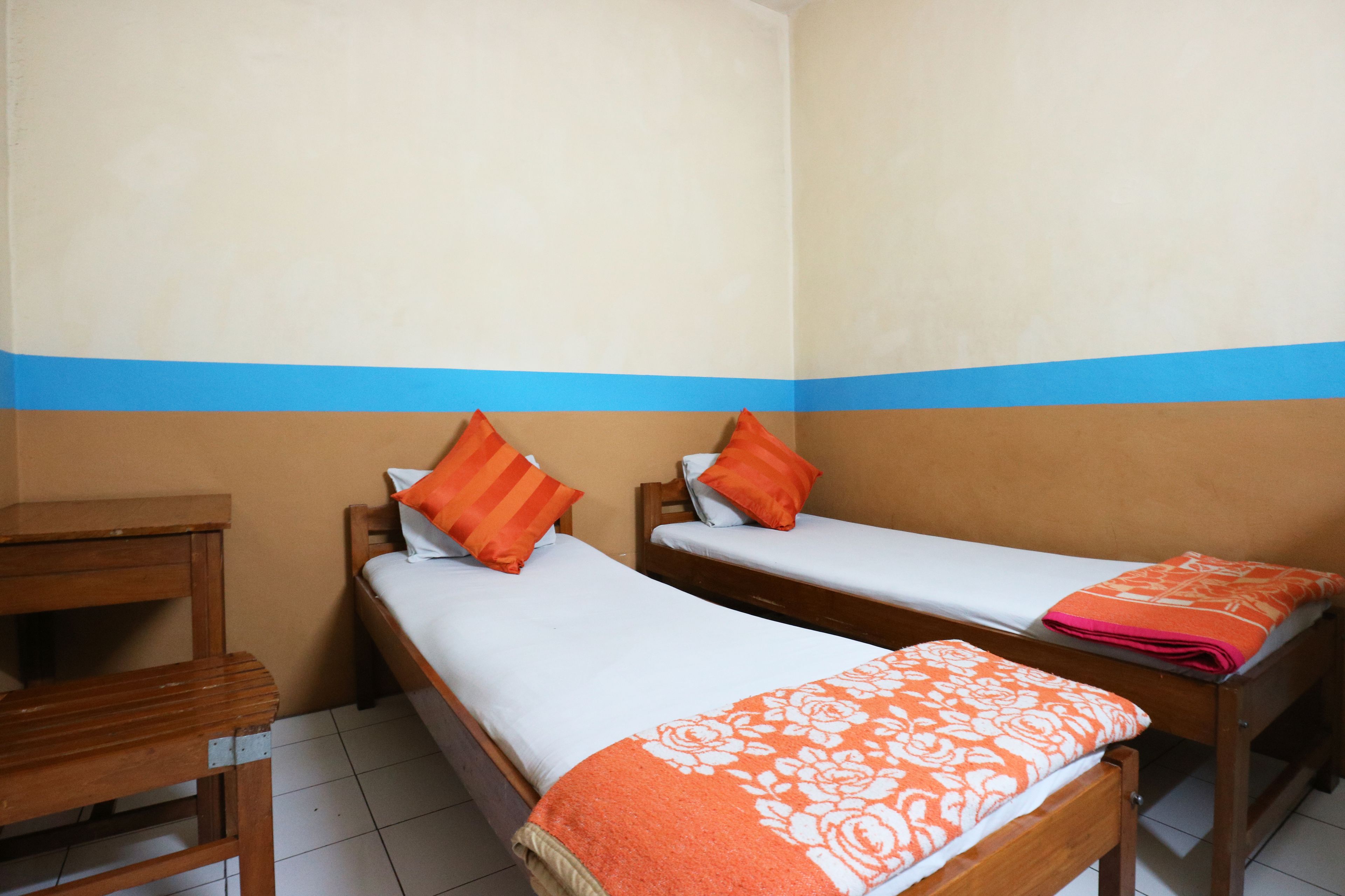 A room with two single beds, each with an orange pillow and a white bedspread. There is a wooden nightstand with a lamp beside one bed. The room has a blue stripe on the wall and a tiled floor.