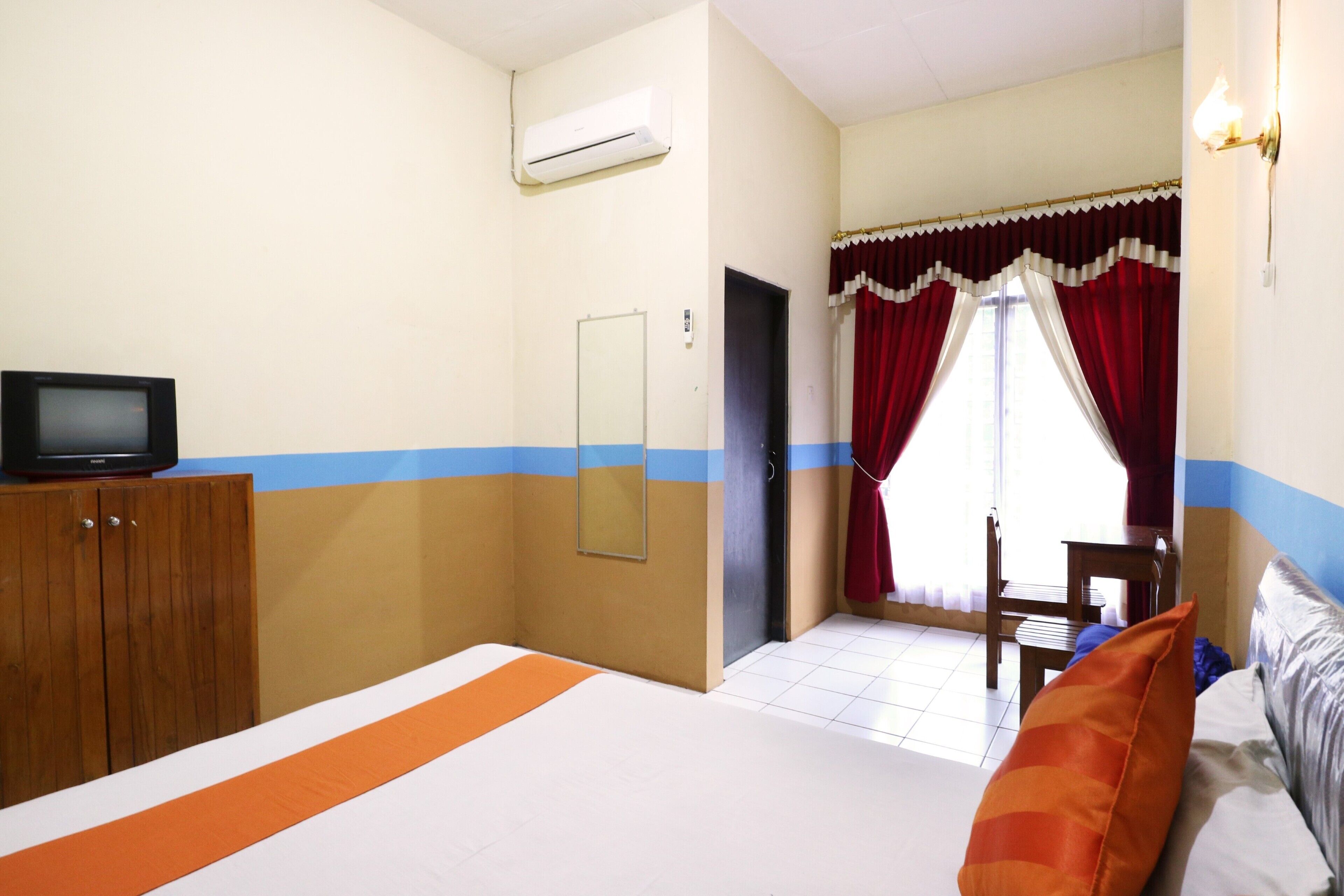 Habitación de hotel con una cama, un televisor sobre un mueble de madera, un espejo, una puerta y una ventana con cortinas rojas.