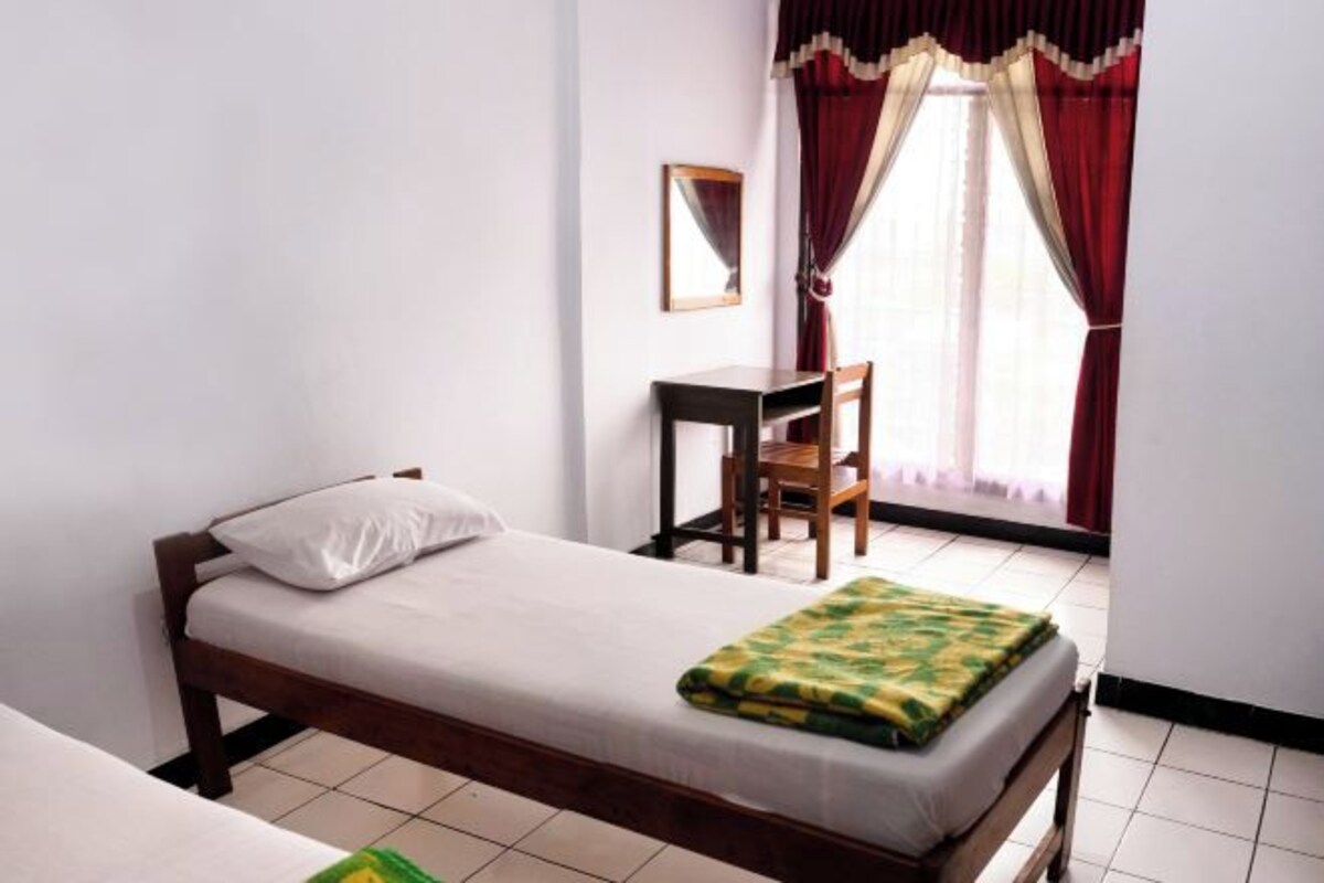 A single bed with a white pillow and a green patterned blanket, a wooden desk with a chair, and a window with red curtains.