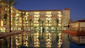 A large hotel with multiple floors, a swimming pool, and palm trees. - Ohtels Islantilla (Lepe)