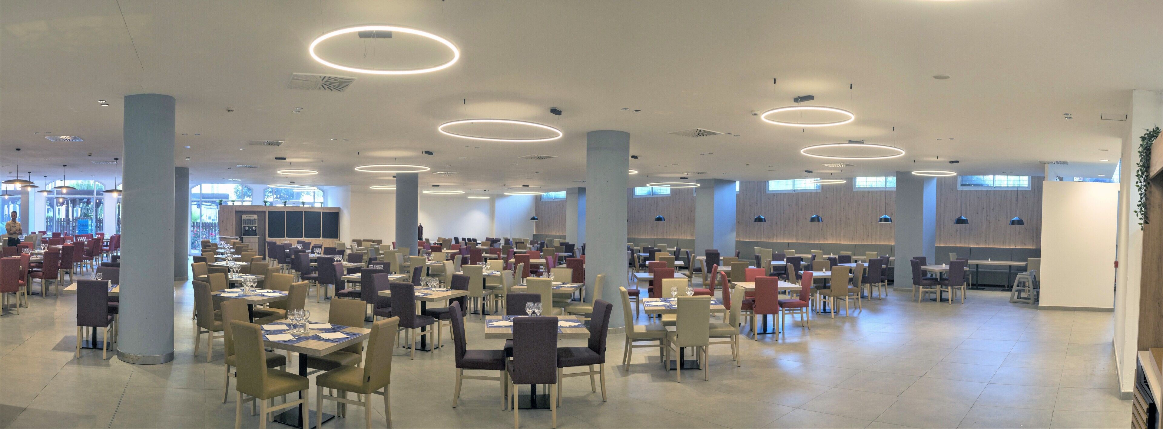 Un comedor amplio con varias mesas y sillas, ventanas grandes y modernas lámparas circulares en el techo.