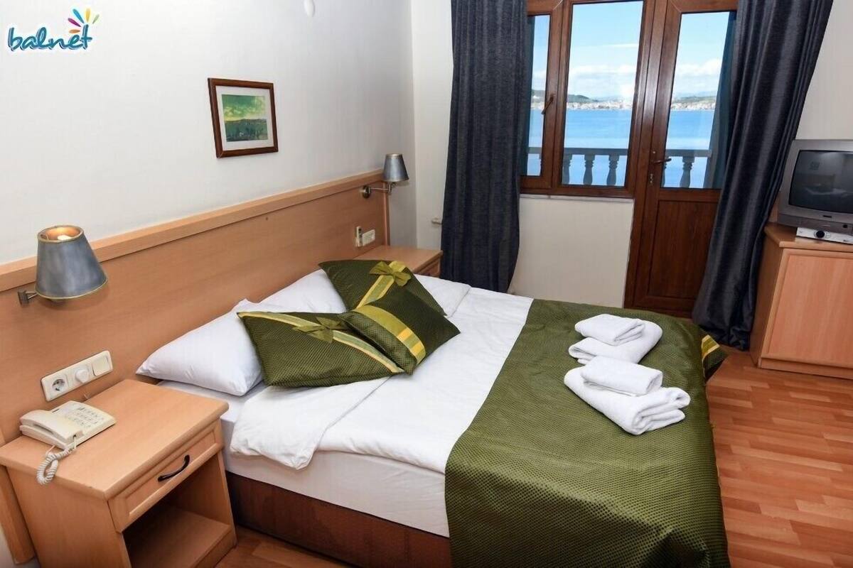 A hotel room with a bed, a TV on a wooden cabinet, a bedside lamp, a telephone, and a window with curtains.