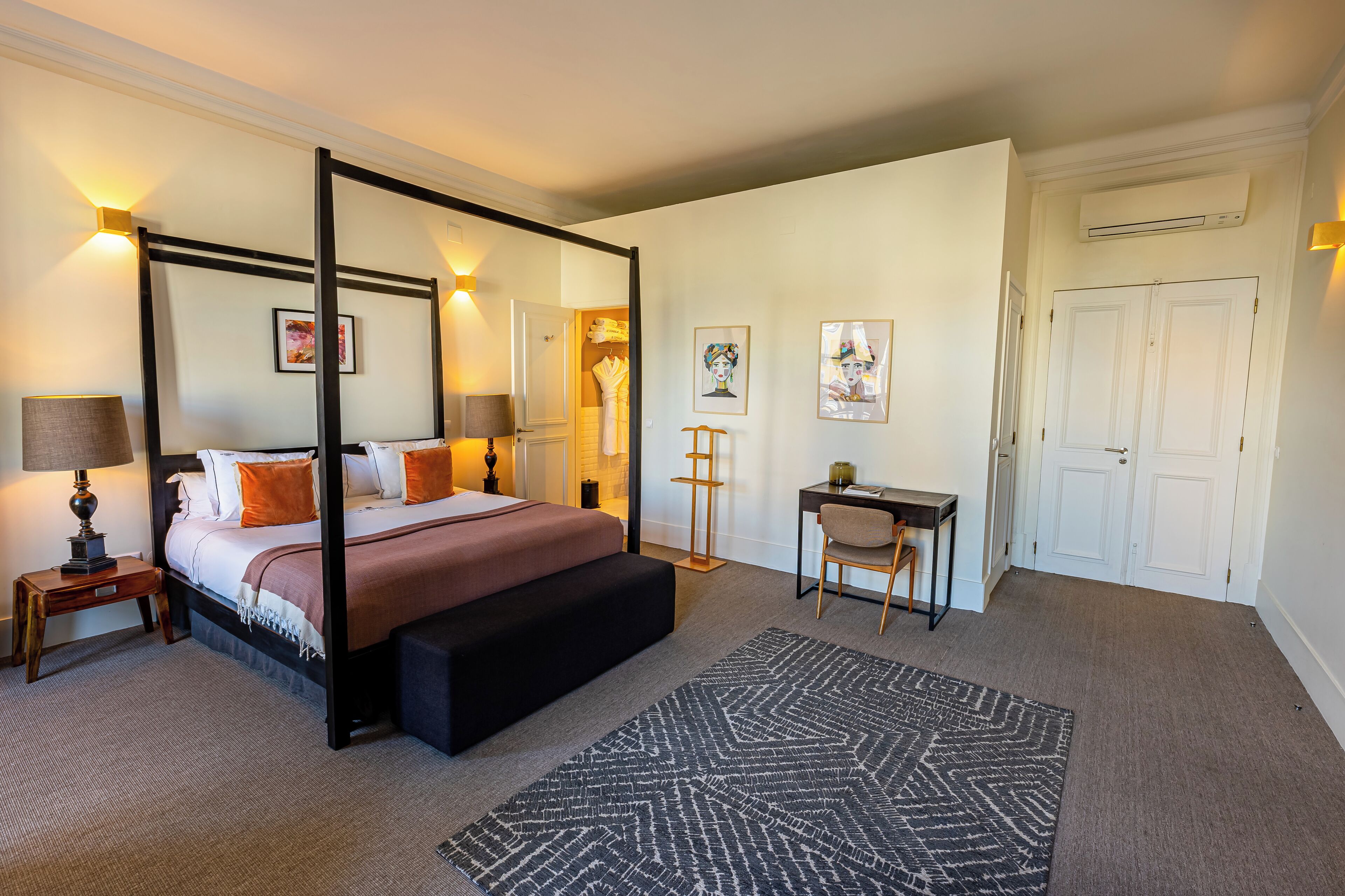 A four-poster bed with a bench, bedside tables, a desk, and chairs in a well-lit room with a carpeted floor.