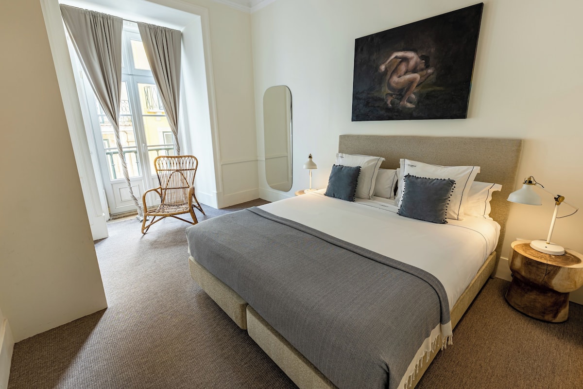 Habitación de hotel con una cama grande, una mesita de noche de madera, una silla, un cuadro en la pared y una ventana con cortinas.