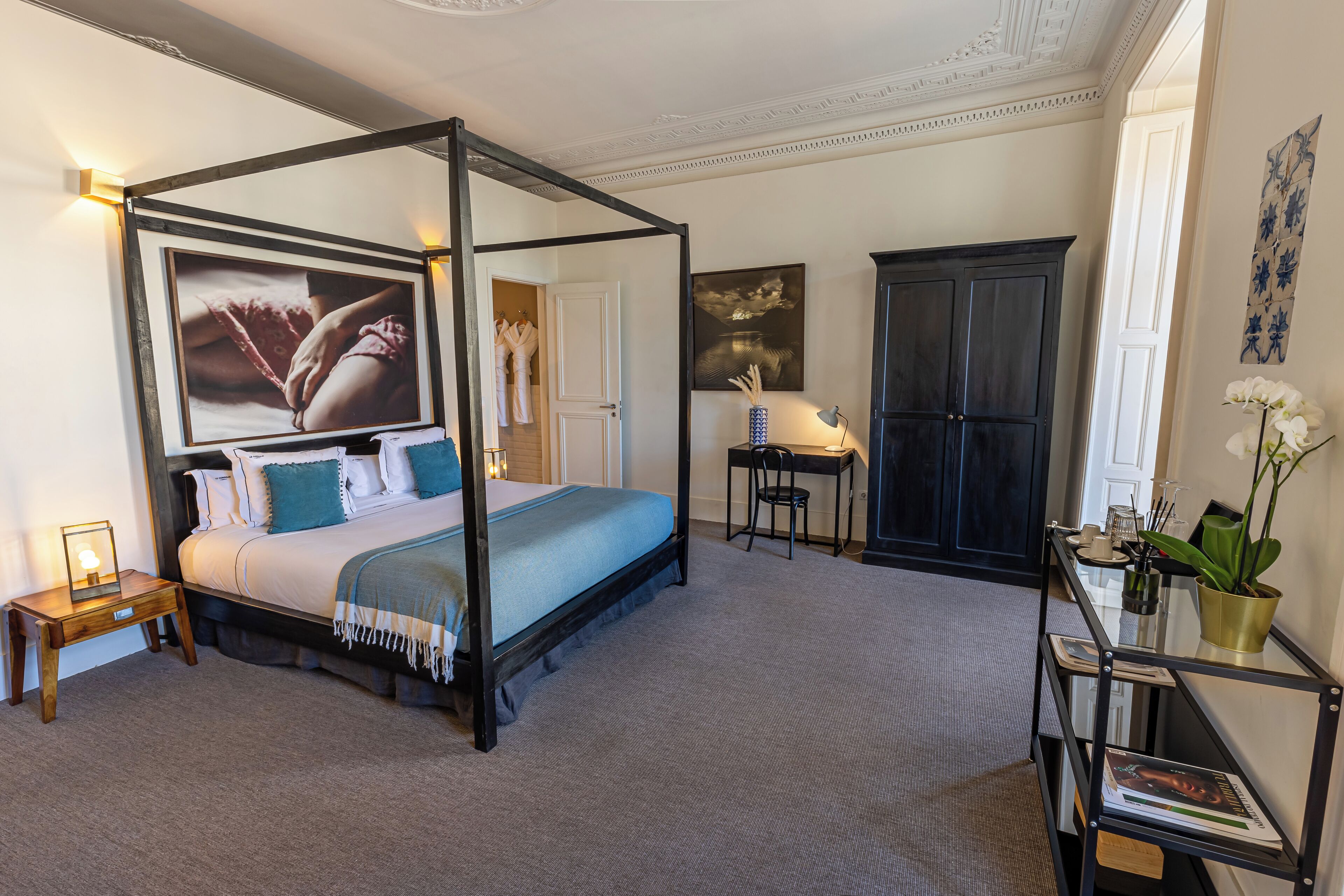A four-poster bed with a blue bedspread, a bedside table with a lamp, a desk with a chair, and a bookshelf with a plant.