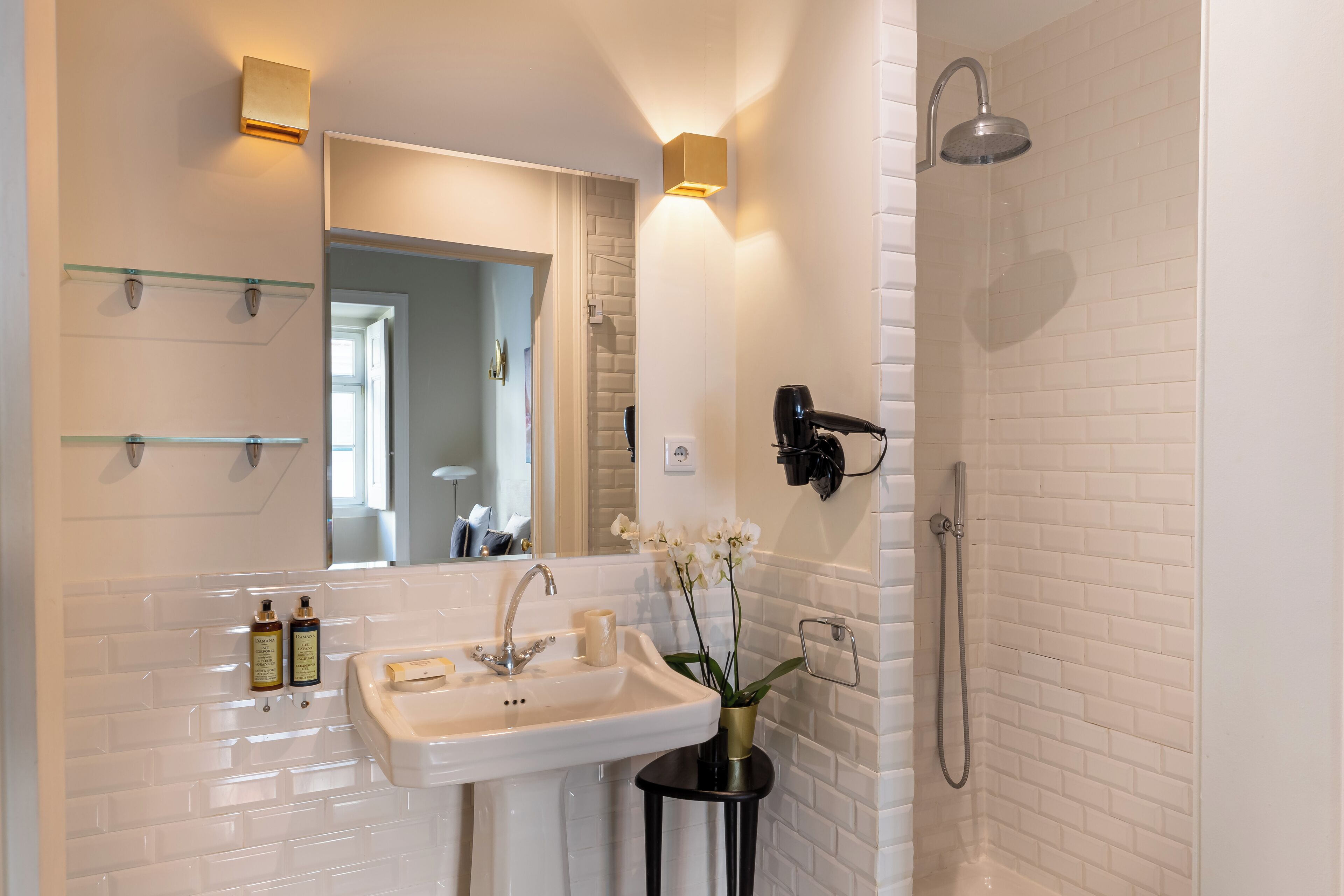 Ein Badezimmer mit weiß gefliesten Wänden, einem Waschbecken mit Spiegel, einer Handbrause und einer Vase mit Blumen.