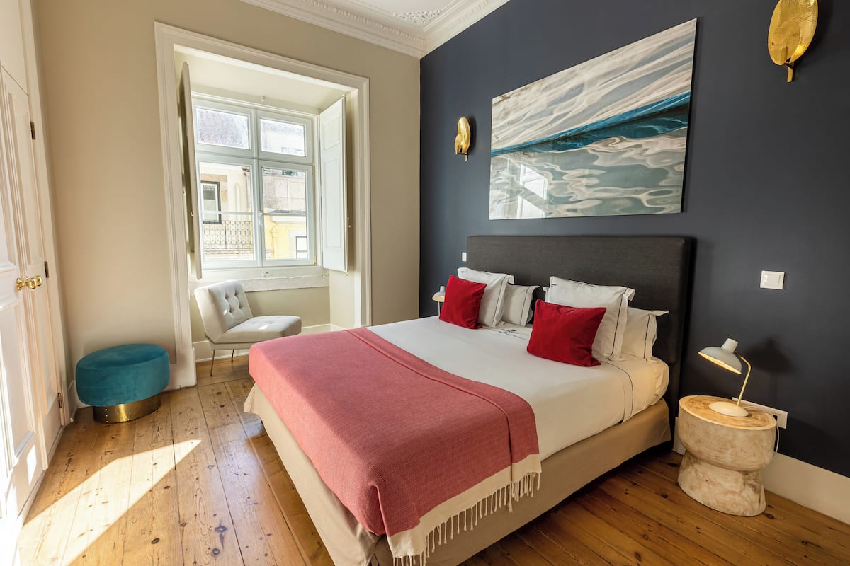 Un dormitorio con cama, ventana, una silla y un cuadro en la pared.