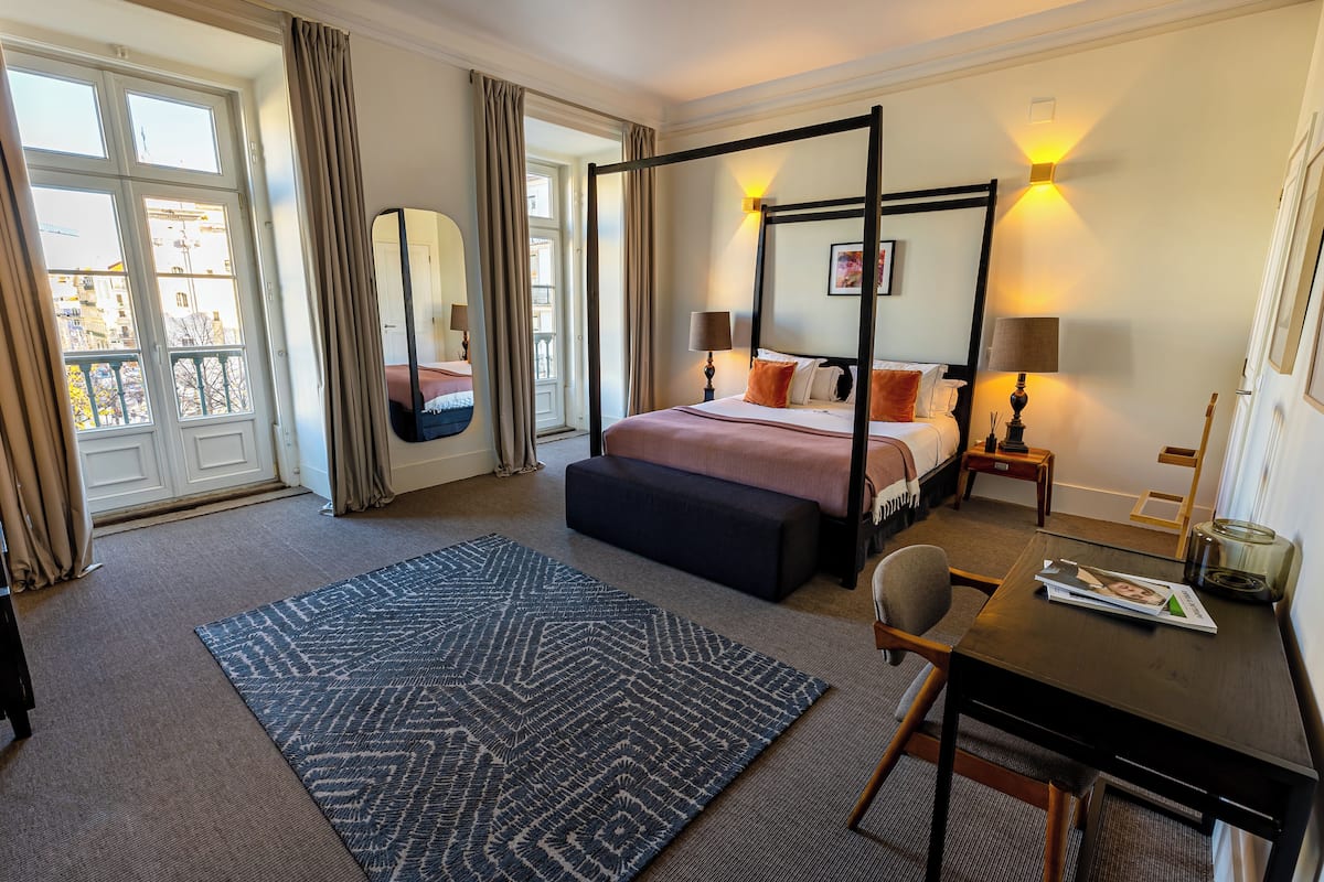 Una cama con dosel, un banco, un escritorio con silla, una alfombra grande y vistas de la ciudad a través de una ventana.
