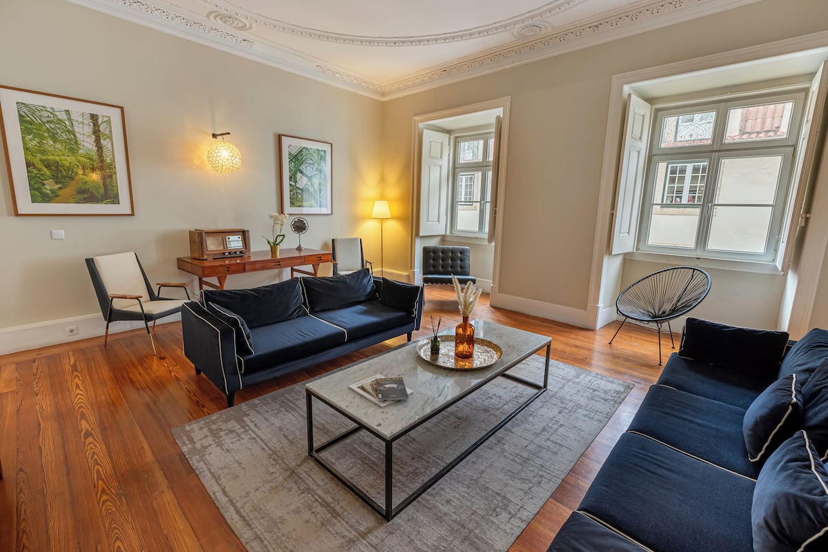 Un salón con un sofá azul oscuro, una mesa de centro de cristal y una mesita auxiliar de madera con una lámpara. Hay dos sillones, una alfombra y un ventanal con vistas a edificios.