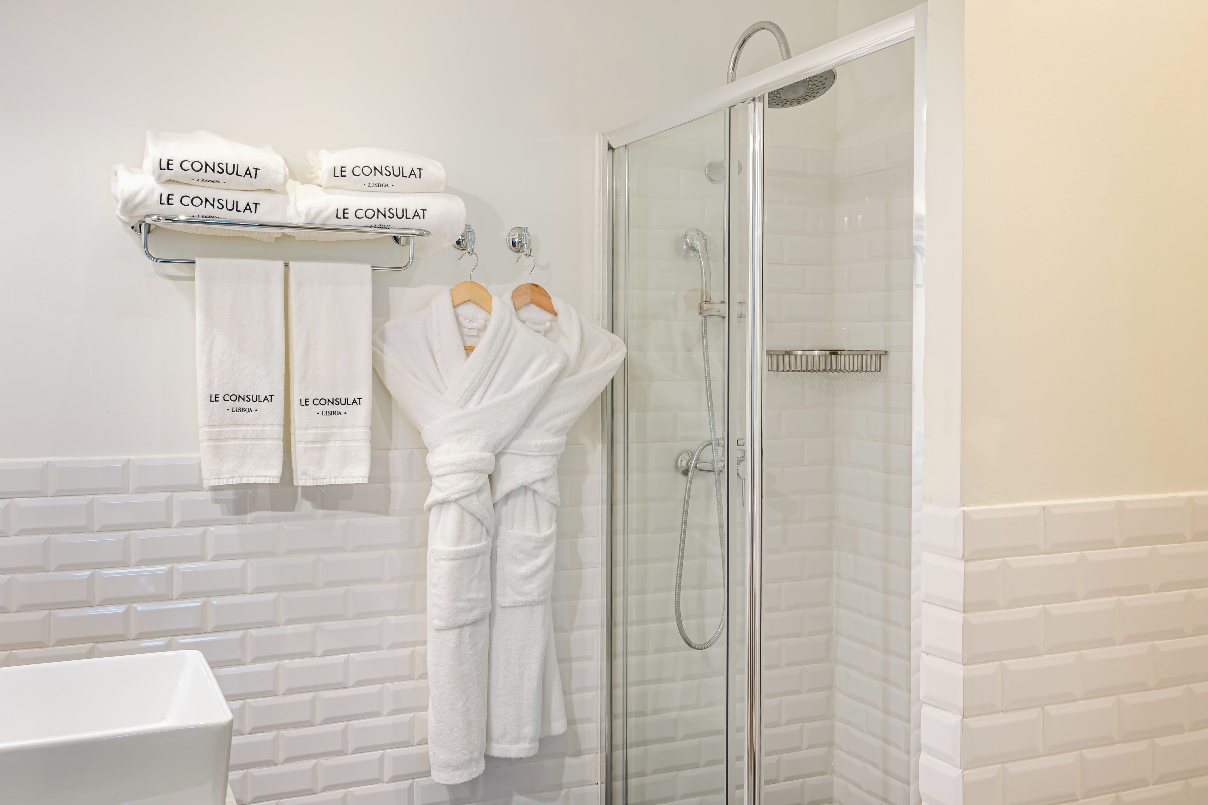 Un baño moderno con cabina de ducha de vidrio, paredes cubiertas de azulejos blancos y un perchero con batas blancas con la etiqueta "LE CONSULAT".