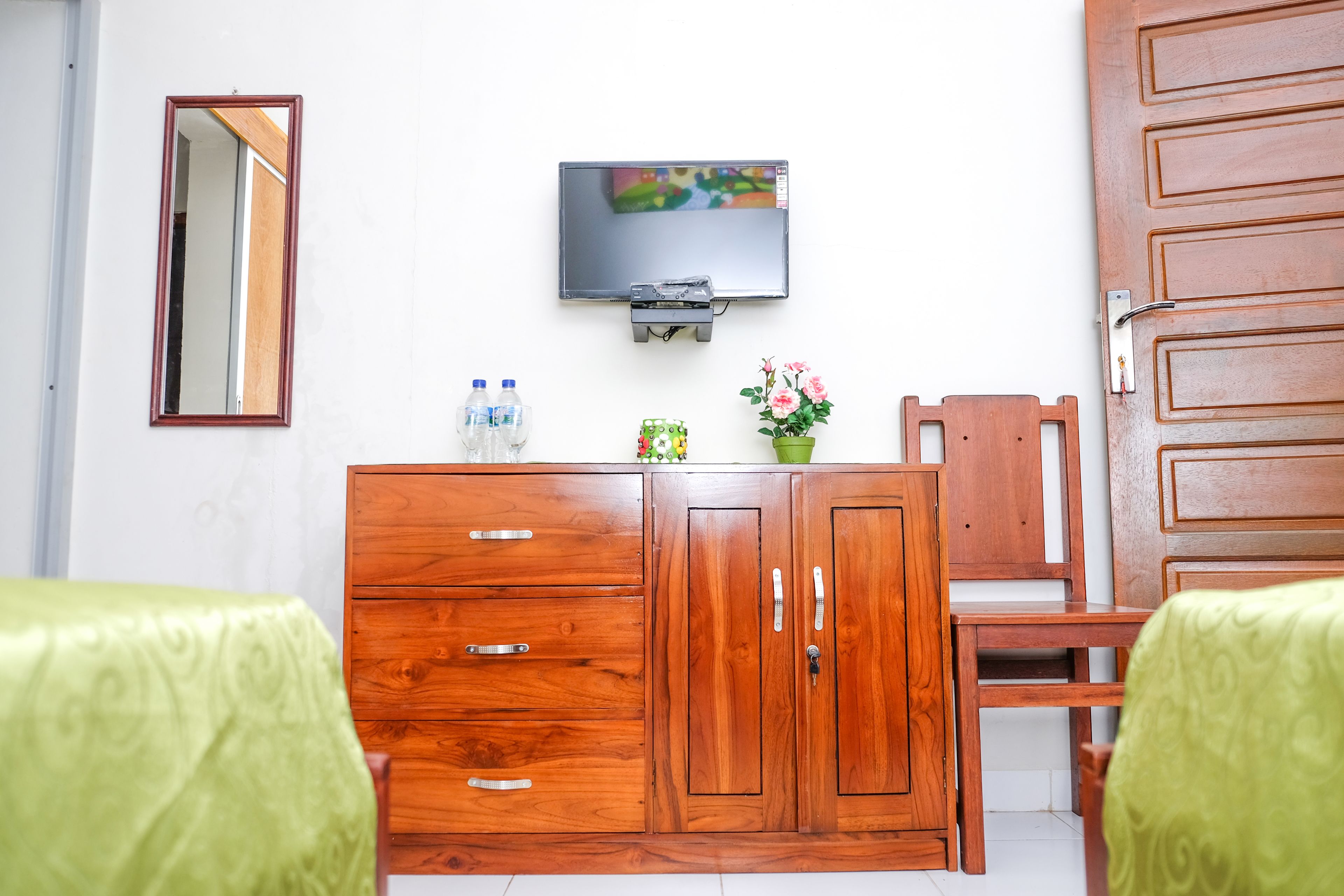 A room with a wooden cabinet, a television mounted on the wall, a chair, and two beds with green covers.