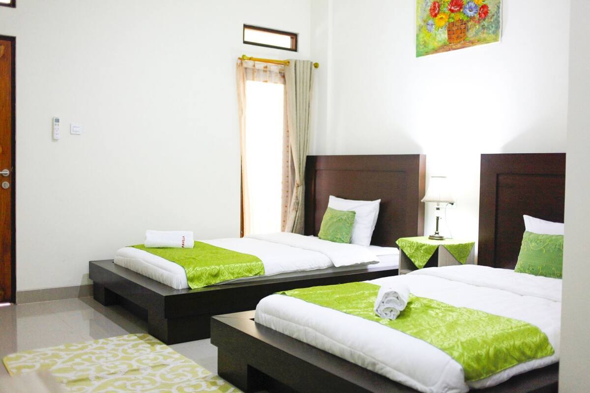 A hotel room with two beds, a small table, a bedside lamp, a window with curtains, and a painting on the wall.