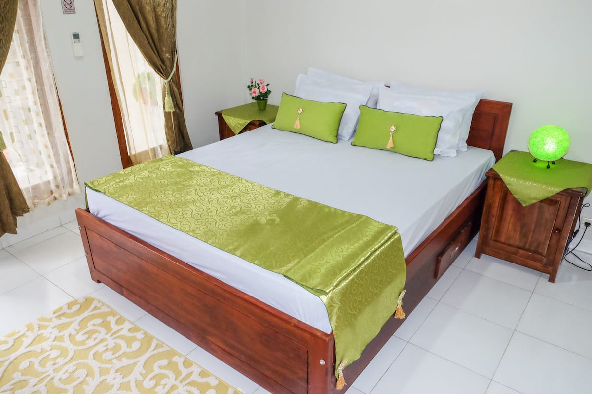 A neatly made bed with a green bedspread and pillows, a bedside table with a green lamp, and a small potted plant on the nightstand.