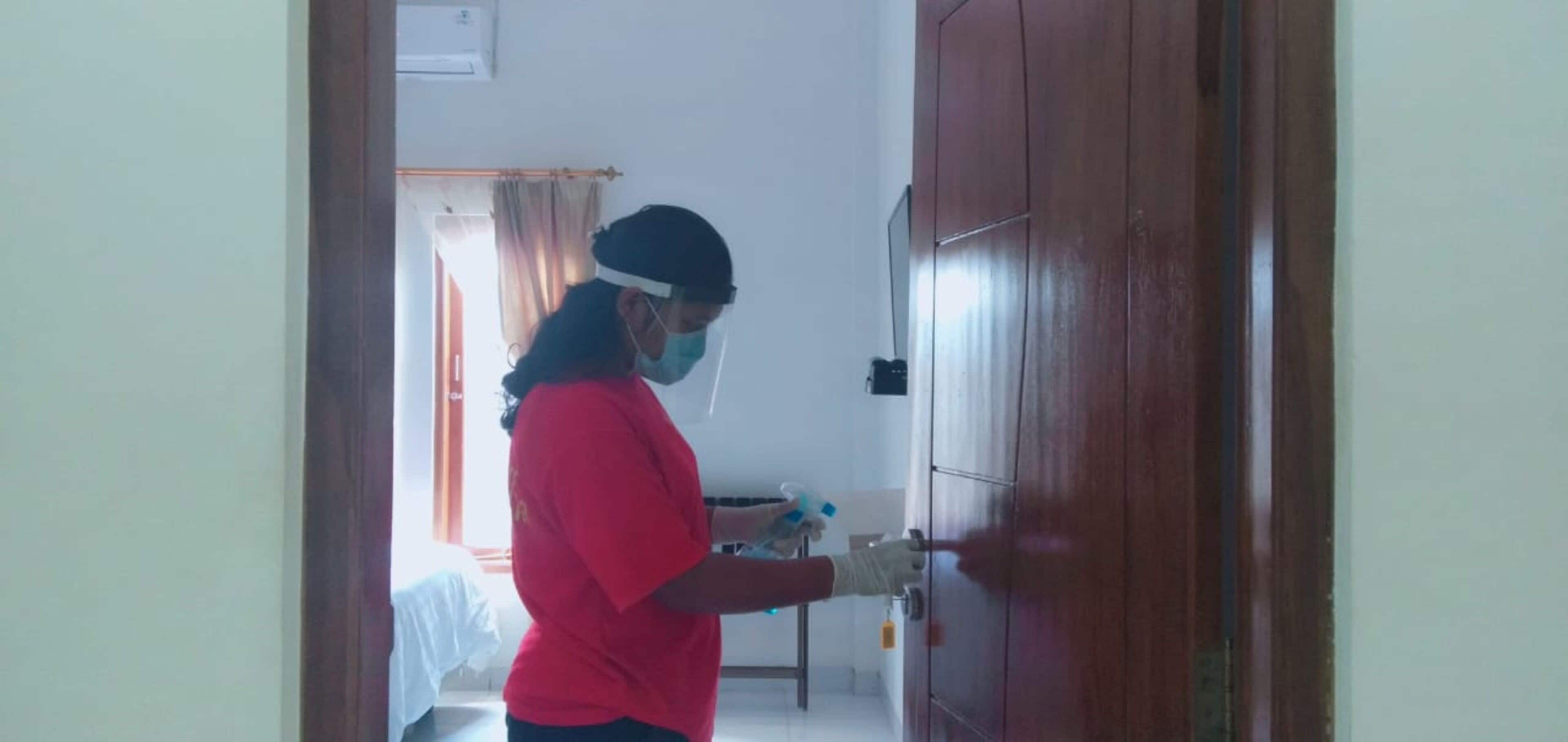 A person wearing a red shirt and a face shield, disinfecting a door handle with a spray bottle.