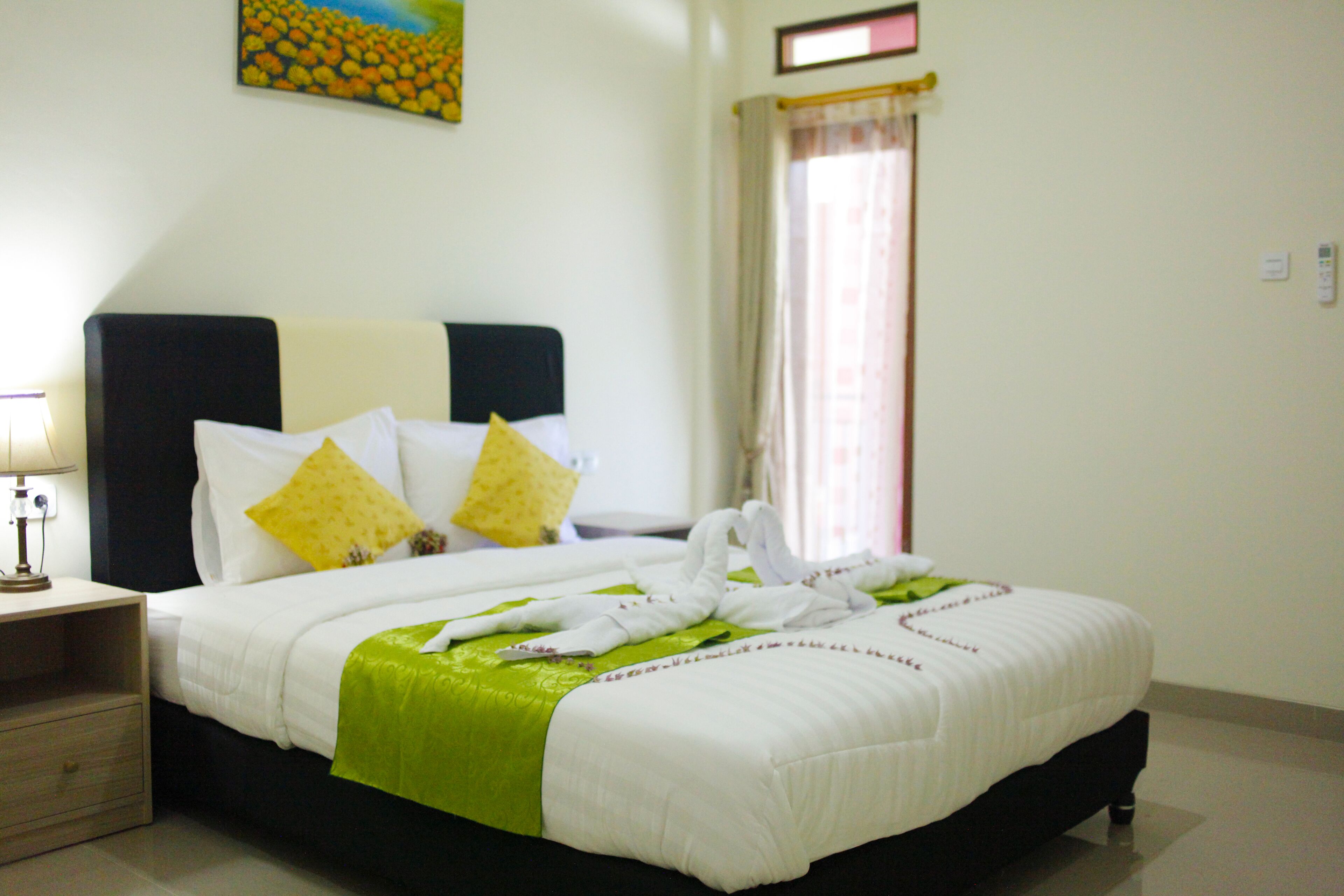 Una habitación de hotel con cama, almohadas, mesita de noche, lámpara, ventana con cortinas y un cuadro en la pared.