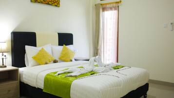 Una habitación de hotel con cama, almohadas, mesita de noche, lámpara, ventana con cortinas y un cuadro en la pared.