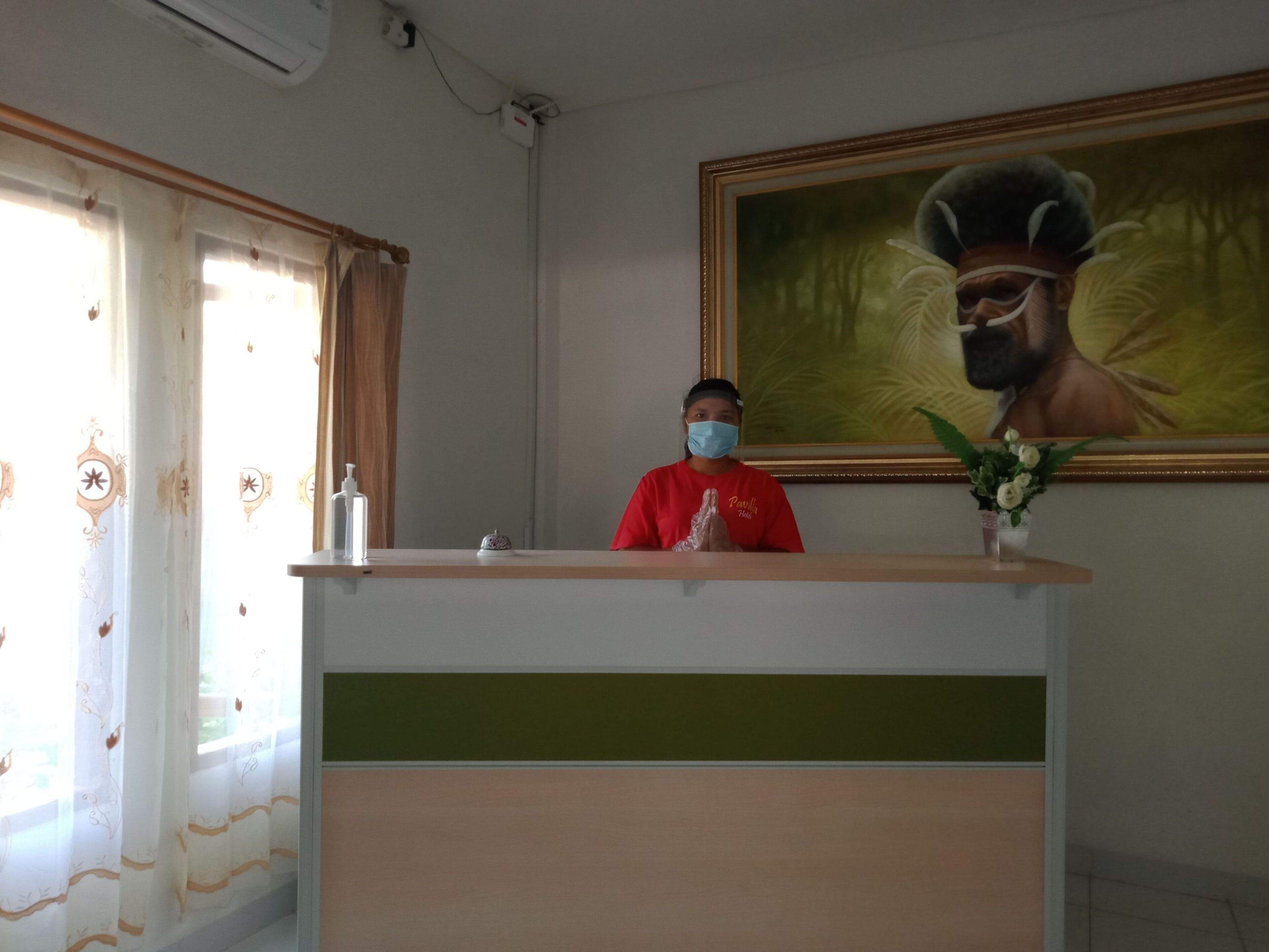 A reception desk with a person in a red shirt, a framed painting on the wall, and a window with curtains.