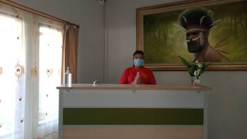 A reception desk with a person in a red shirt, a framed painting on the wall, and a window with curtains.