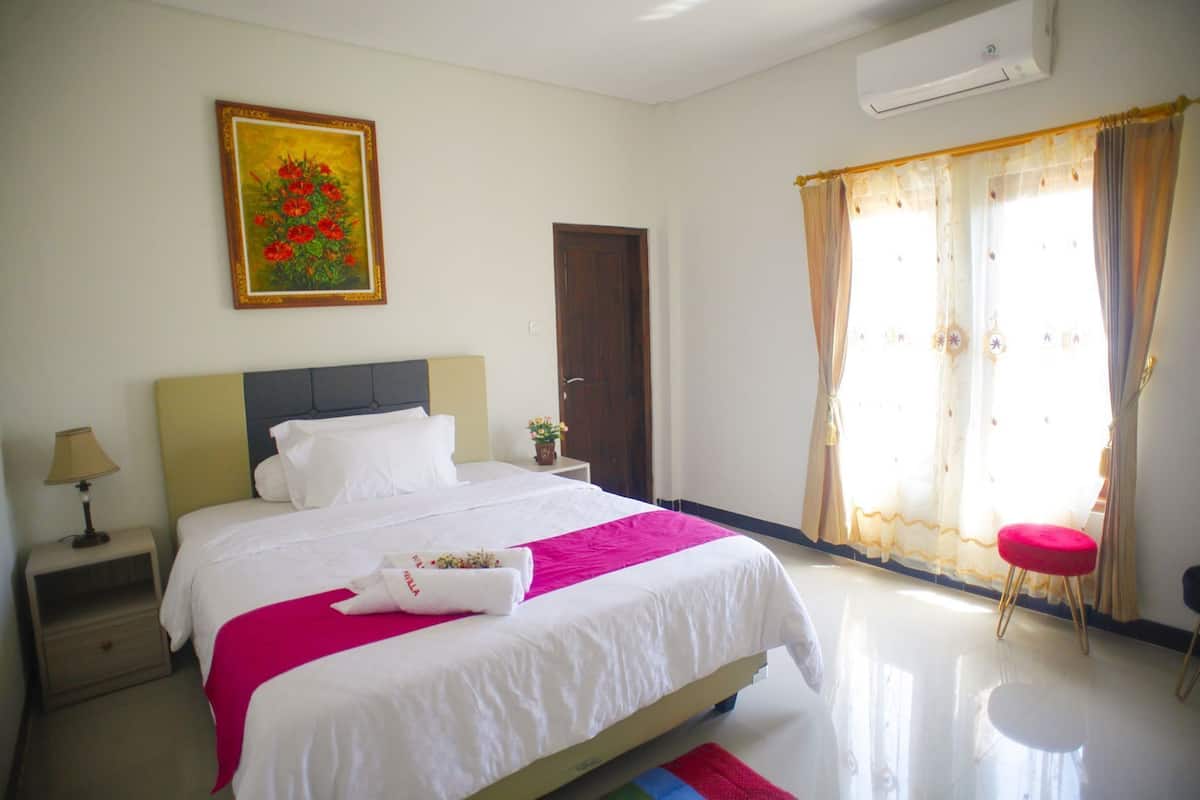 A hotel room with a bed, a nightstand, a lamp, a painting, a door, and a window with curtains.