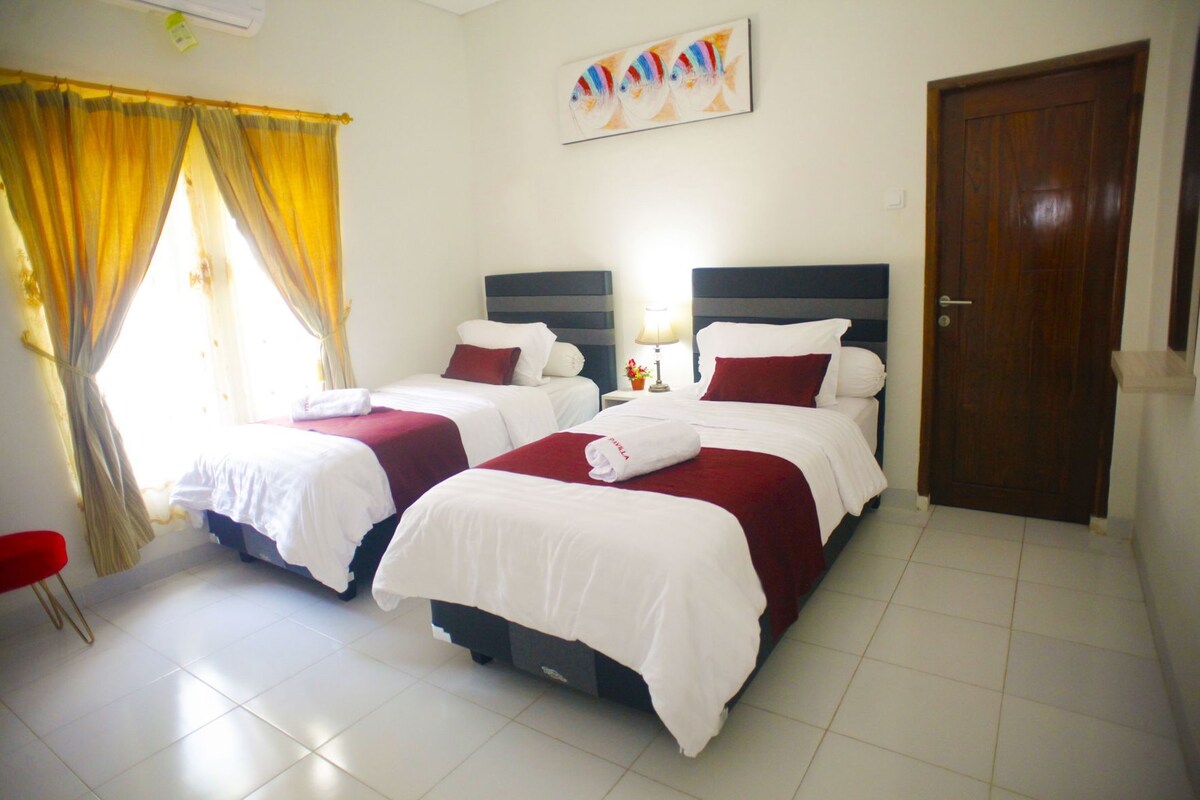 A hotel room with two beds, a door, a window with curtains, a bedside table with a lamp, and a painting on the wall.
