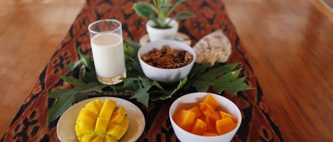Ein Tisch mit einem gemusterten Tischtuch, ein Glas Milch, eine Schüssel Granola, eine Schüssel mit Mangostücken und eine weitere mit Mangowürfeln, eine Topfpflanze und ein Stück Brot.