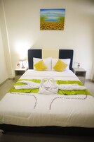 A neatly made bed with white bedding and two yellow pillows, a painting of a landscape on the wall, and a green throw blanket with towels on the bed.