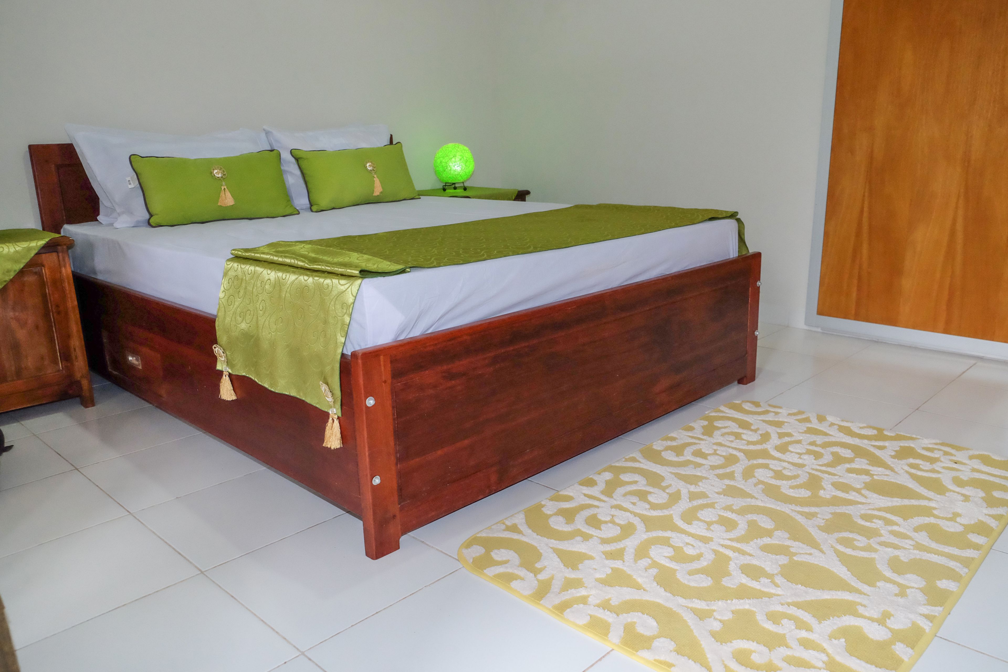 Una cama bien tendida con ropa de cama verde y blanca, un cabecero de madera y una alfombra estampada amarilla sobre piso de baldosas.
