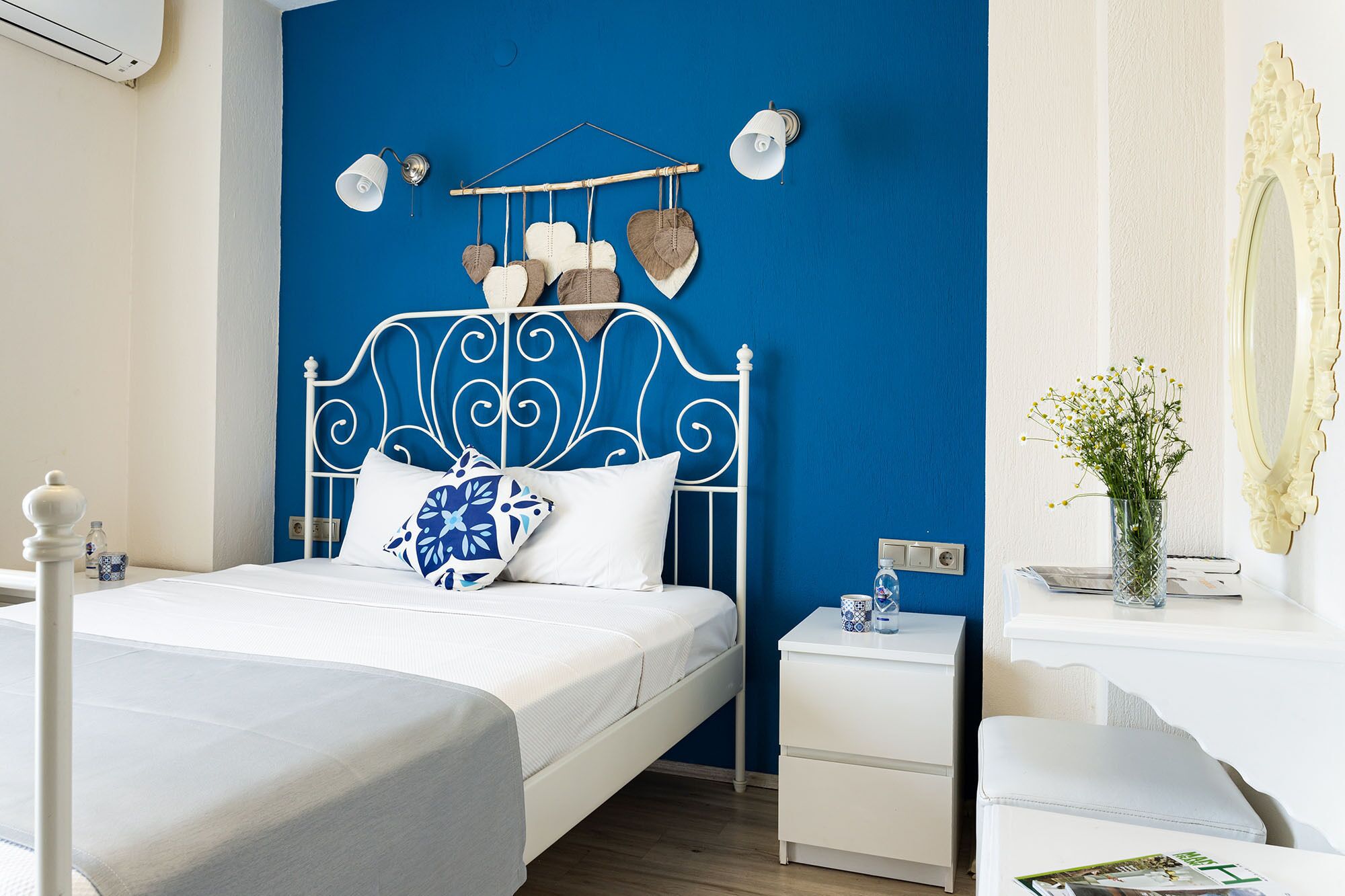 Ein Schlafzimmer mit blauer Akzentwand, einem weißen Metallbettgestell, einem Nachttisch mit Lampe und einem Beistelltisch mit Blumenvase.